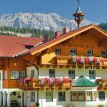 Ein großes Holzhaus im alpinen Stil mit Blumenkästen auf den Balkonen, umgeben von Bäumen und Bergen im Hintergrund unter einem klaren Himmel.