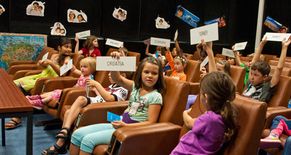 Die Kinder sitzen auf Stühlen im Hörsaal und halten Länderkarten mit Namen wie Kroatien, Türkei und Dänemark hoch, während sie scheinbar eine pädagogische Aktivität durchführen.