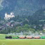 Ein Dampfzug fährt durch eine Wiese, im Hintergrund sind ein Dorf, bewaldete Hügel und eine Burg zu sehen.