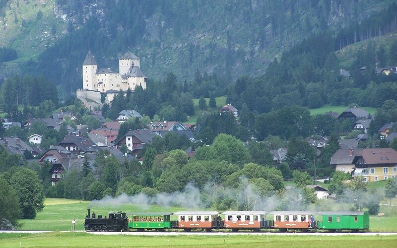Ein Dampfzug fährt durch eine Wiese, im Hintergrund sind ein Dorf, bewaldete Hügel und eine Burg zu sehen.