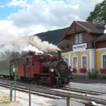 Ein roter Dampfzug kommt an einem kleinen Bahnhof mit der Aufschrift „Mauterndorf“ an. In der Nähe stehen Passagiere und eine Frau mit zwei Kindern und schauen zu.