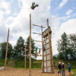Eine Gruppe von Menschen nimmt an einem Hochseilgarten im Freien teil; eine Person hängt kopfüber in der Luft über der Struktur, während die anderen vom Boden aus zuschauen.