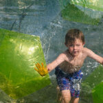 Ein kleiner Junge in Badehose läuft in einem großen aufblasbaren Plastikball auf dem Wasser und drückt seine Hände gegen die transparente Oberfläche.