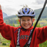 Lächelndes Kind mit Helm und rotem Pullover hält sich im Freien an Seilen fest, im Hintergrund grüne Hügel und Berge.