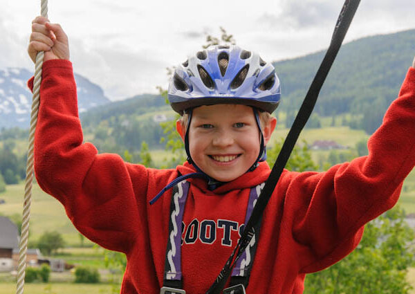 Lächelndes Kind mit Helm und rotem Pullover hält sich im Freien an Seilen fest, im Hintergrund grüne Hügel und Berge.