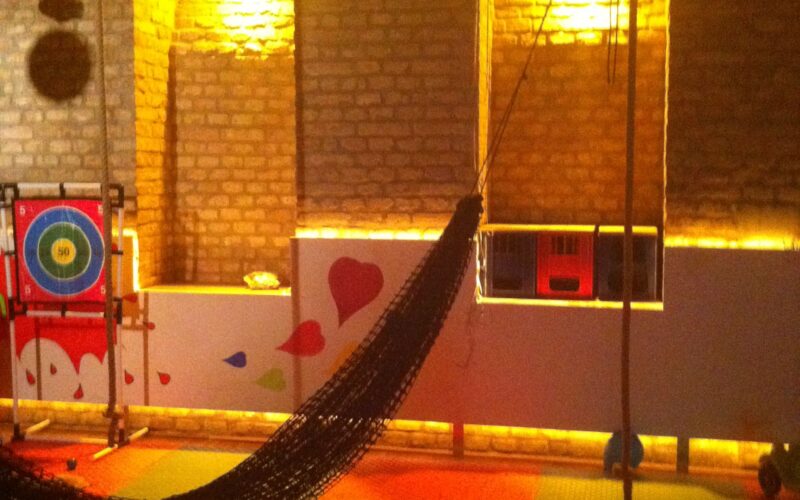 Innenspielbereich mit Backsteinwänden, weichem, farbenfrohem Bodenbelag, einem hängenden Netz, runden Schaumstoffblöcken und einem Zielbrett, beleuchtet von warmem, gelbem Licht.