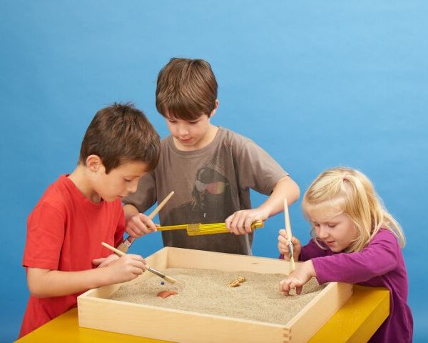 Vor einem schlichten blauen Hintergrund graben drei Kinder mit Werkzeugen in einer mit Sand gefüllten Tischkiste und suchen nach Gegenständen.