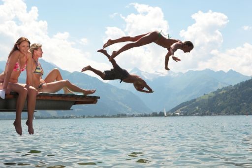 Zwei Personen sitzen auf einem Steg, während zwei andere in einen See springen, mit Bergen und einem bewölkten Himmel im Hintergrund.