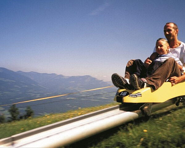 Ein Erwachsener und ein Kind fahren mit einer gelben Alpenachterbahn einen Berg hinunter, mit malerischen Hügeln und einem klaren Himmel im Hintergrund.
