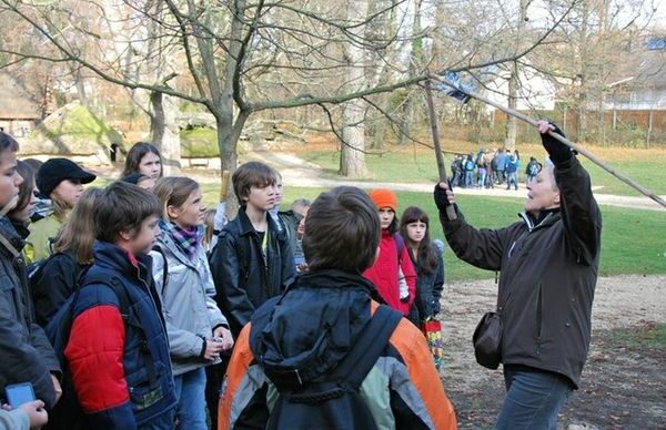 Eine Gruppe Kinder steht draußen in einem Park und hört einem Erwachsenen zu, der einen langen Stock hält und beim Sprechen gestikuliert.