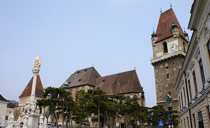 Historische Steingebäude mit einem hohen Uhrenturm, einem kunstvollen Denkmal und Bäumen davor, unter einem klaren Himmel.