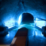 Schwach beleuchteter unterirdischer Tunnel mit blauer Beleuchtung, durch die Mitte fließendem Wasser und einer gewölbten Decke.