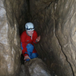 Eine Person mit Helm und roter Jacke hockt in einem engen, von Felswänden umgebenen Höhlengang.