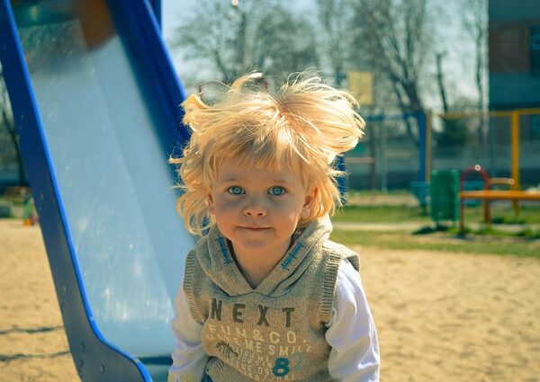 Ein kleines Kind mit blonden Haaren steht an einem sonnigen Tag neben einer blauen Rutsche auf einem Spielplatz, im Hintergrund sind Sand und Spielgeräte zu sehen.