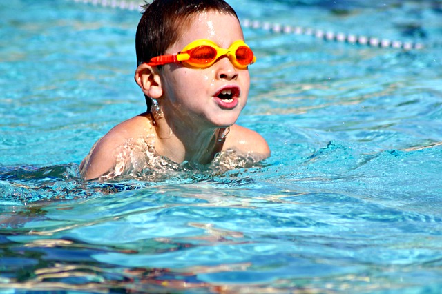 Ein kleiner Junge mit einer orangefarbenen Schwimmbrille schwimmt in einem Pool. Um ihn herum spritzt Wasser, im Hintergrund ist eine Bahnbegrenzung zu sehen.