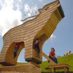 Große Holzkonstruktion in Form eines stilisierten Tieres, unter der Kinder klettern und spielen, auf einem grasbewachsenen Hügel unter einem teilweise bewölkten Himmel.