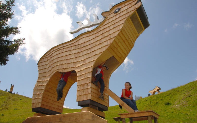 Große Holzkonstruktion in Form eines stilisierten Tieres, unter der Kinder klettern und spielen, auf einem grasbewachsenen Hügel unter einem teilweise bewölkten Himmel.