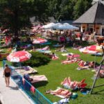 Menschen, die sich an einem sonnigen Tag auf einer Wiese am Seeufer mit Sonnenschirmen, einer Spielplatzschaukel und einem kleinen Gebäude in der Nähe sonnen und entspannen.