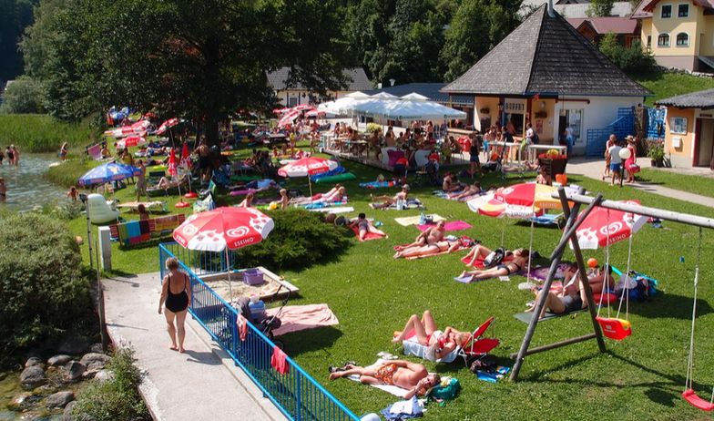 Menschen, die sich an einem sonnigen Tag auf einer Wiese am Seeufer mit Sonnenschirmen, einer Spielplatzschaukel und einem kleinen Gebäude in der Nähe sonnen und entspannen.