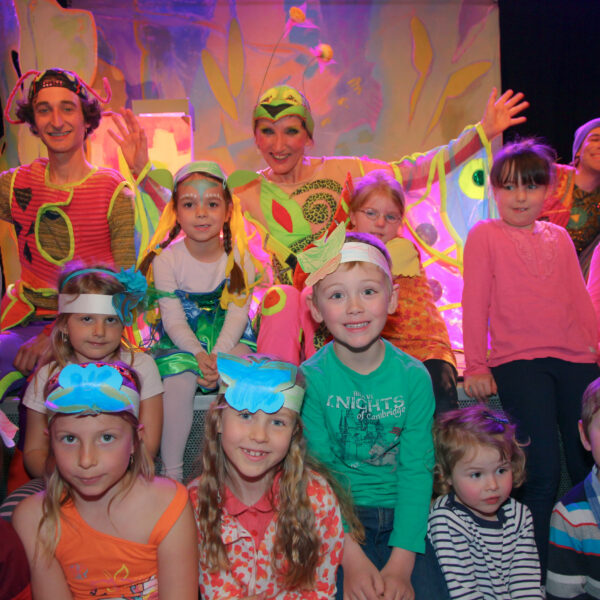 Kinder und zwei Erwachsene in bunten Kostümen sitzen und posieren für ein Gruppenfoto auf der Bühne, mit einer bunten Kulisse im Hintergrund.