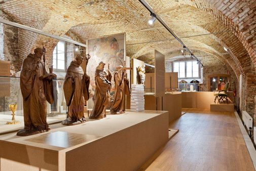 In einer Museumsgalerie mit Ziegelgewölbedecken werden mehrere lebensgroße Statuen und Artefakte in Glasvitrinen ausgestellt, die von Deckenscheinwerfern beleuchtet werden.