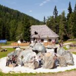 Die Menschen sind um und auf großen, kreisförmig angeordneten Steinen versammelt, mit einer Holzhütte und Kiefern im Hintergrund an einem sonnigen Tag.