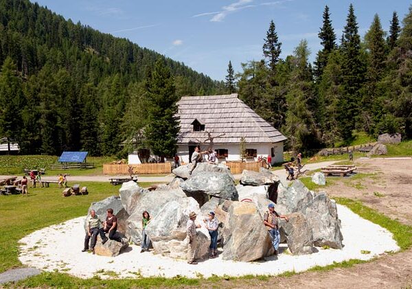 Die Menschen sind um und auf großen, kreisförmig angeordneten Steinen versammelt, mit einer Holzhütte und Kiefern im Hintergrund an einem sonnigen Tag.