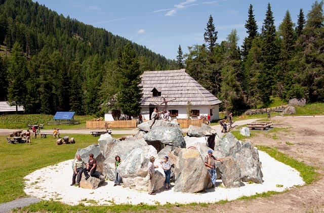 Die Menschen sind um und auf großen, kreisförmig angeordneten Steinen versammelt, mit einer Holzhütte und Kiefern im Hintergrund an einem sonnigen Tag.
