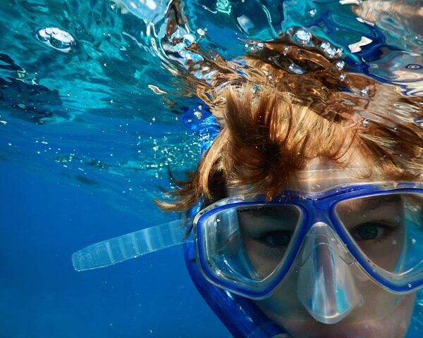 Eine Person mit Schnorchelmaske befindet sich teilweise unter Wasser, wobei ihr Gesicht und ihre Haare durch das klare blaue Wasser sichtbar sind.