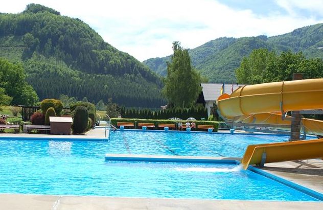 Außenpool mit gelber Wasserrutsche, umgeben von Grün und Bergen im Hintergrund unter einem teilweise bewölkten Himmel.