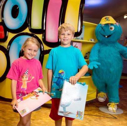 Zwei Kinder halten bunte Notizbücher und stehen vor einer großen kostümierten blauen Figur und einem farbenfrohen Display mit der Aufschrift „Lolli Pop“ im Hintergrund.