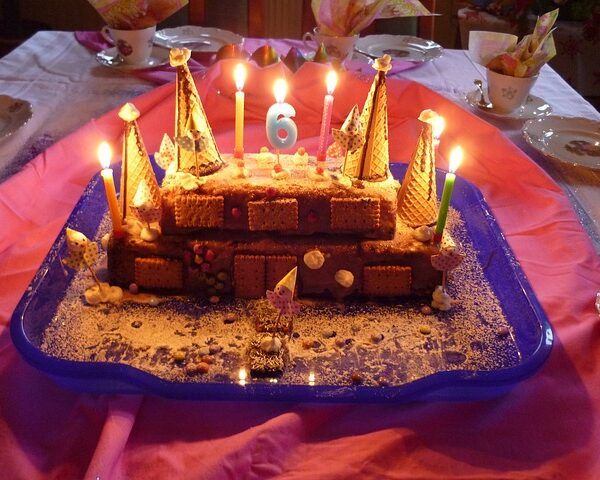 Auf einem blauen Tablett steht eine schlossförmige Geburtstagstorte mit brennenden Kerzen und einer blauen Zahl 6 obendrauf, dekoriert mit Oblaten und Keksen.