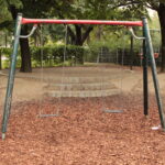 Eine leere Schaukel mit zwei Sitzen steht auf Holzspänen in einem Park, umgeben von Bäumen und einem eingezäunten Gelände im Hintergrund.