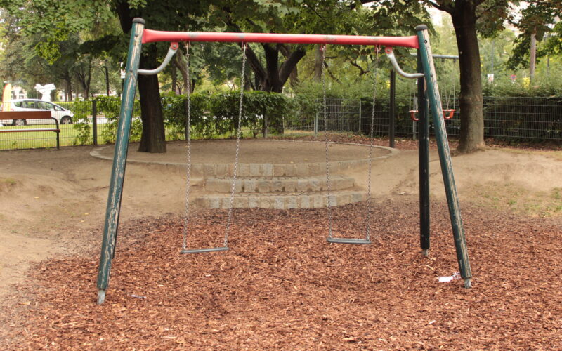 Eine leere Schaukel mit zwei Sitzen steht auf Holzspänen in einem Park, umgeben von Bäumen und einem eingezäunten Gelände im Hintergrund.