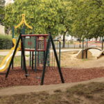 Ein Spielplatz mit einer gelben Rutsche, einem Klettergerüst und einer Schaukel liegt auf Mulch, neben einer kleinen Holzbrücke und einer grünen Federwippe, umgeben von Bäumen.