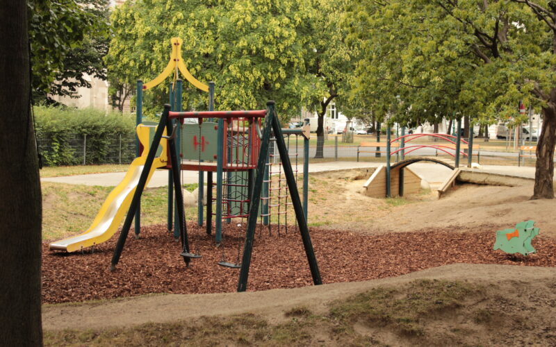 Ein Spielplatz mit einer gelben Rutsche, einem Klettergerüst und einer Schaukel liegt auf Mulch, neben einer kleinen Holzbrücke und einer grünen Federwippe, umgeben von Bäumen.