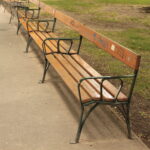 Eine Reihe leerer Holzbänke mit Metallrahmen und einigen Graffiti auf den Rückenlehnen säumen einen gepflasterten Weg in einem Park mit Gras und einem Baum in der Nähe.