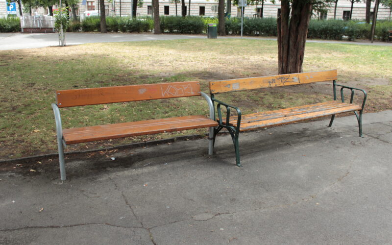 Zwei hölzerne Parkbänke, eine intakt und eine beschädigt, bei der Latten fehlen, stehen nebeneinander auf einer gepflasterten Fläche, umgeben von Gras und Bäumen.