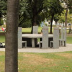 Ein runder Steintisch mit sechs passenden Stühlen steht auf einer kreisförmigen, gepflasterten Fläche in einem Park, umgeben von Gras und Bäumen. Ein Baumstamm verdeckt teilweise die linke Bildseite.