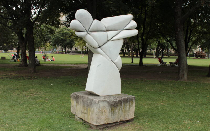 Abstrakte Steinskulptur mit geschwungenen, sich kreuzenden Formen auf einem Betonsockel, aufgestellt auf einer Rasenfläche in einem Park mit Bäumen und Menschen im Hintergrund.