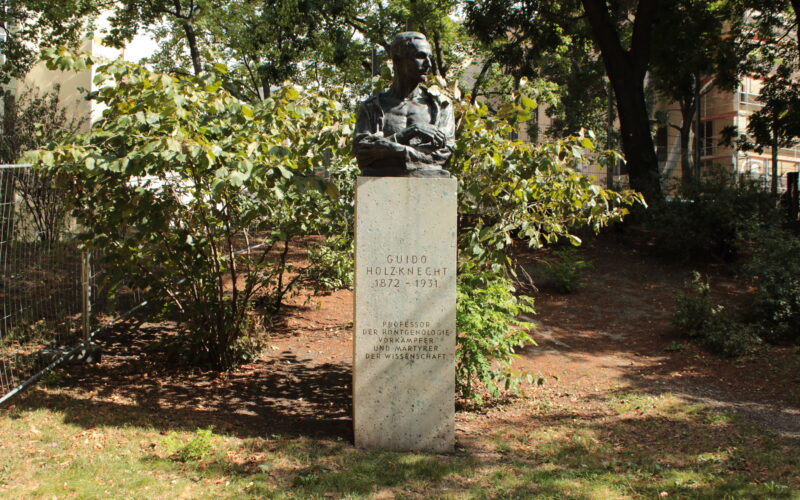 Eine Bronzebüste von Guido Holzknecht auf einem Steinsockel in einem Park mit Bäumen und Büschen im Hintergrund. Auf dem Sockel sind sein Name, sein Lebensjahr und seine Leistungen eingraviert.