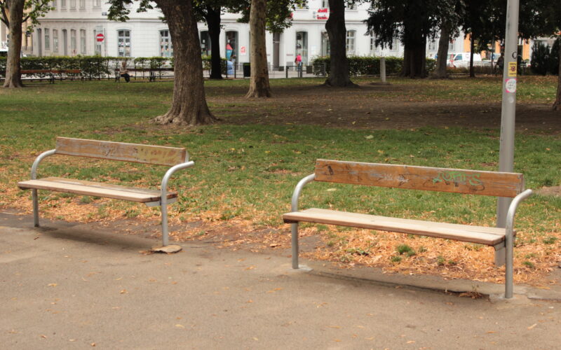 Zwei leere Holzbänke mit Metallrahmen stehen auf einem gepflasterten Weg in einem Park, umgeben von Gras, Bäumen und einigen verstreuten Blättern.