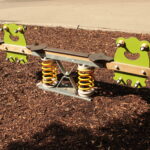 Eine Spielplatzwippe mit froschförmigen Sitzen und gelben Federn ist auf einer Mulchfläche aufgestellt.