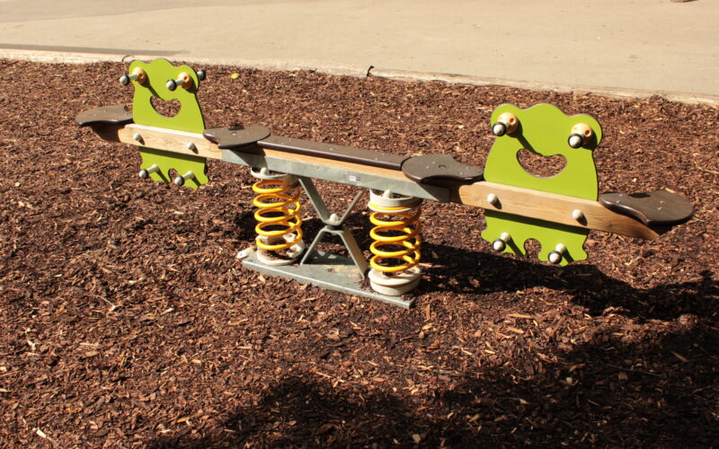Eine Spielplatzwippe mit froschförmigen Sitzen und gelben Federn ist auf einer Mulchfläche aufgestellt.