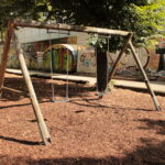 Holzschaukel mit zwei Schaukeln auf einem mit Holzspänen bedeckten Spielplatz, mit Bäumen und einer mit Graffiti bedeckten Mauer im Hintergrund.