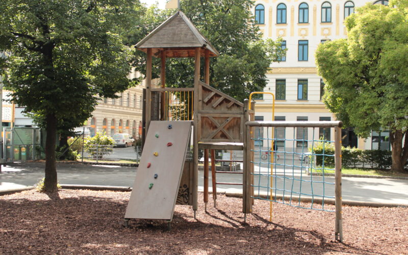 Ein hölzernes Spielgerät mit Rutsche, Kletterwand, Seilnetz und Leiter steht auf Rindenmulch in einem von Bäumen und Gebäuden umgebenen Stadtpark.