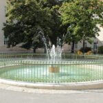 Ein kleiner runder Brunnen mit Wasserstrahlen ist von einem grünen Metallzaun umgeben und befindet sich in einem gepflasterten Innenhof mit Bäumen und Gebäuden im Hintergrund.