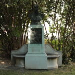 Bronzenes Büstendenkmal mit gravierter Gedenktafel und zwei geschwungenen Bänken, umgeben von Laubbäumen in einer Gartenanlage.