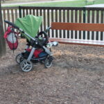 Ein Kinderwagen mit grünem Vordach wird neben einer Holzbank in einem Park abgestellt. Am Griff des Kinderwagens hängt eine Netztasche.