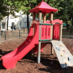 Rote Spielplatzstruktur mit Rutsche und Kletterwand, umgeben von einem Holzzaun und Mulch, mit Bäumen und Gebäuden im Hintergrund.